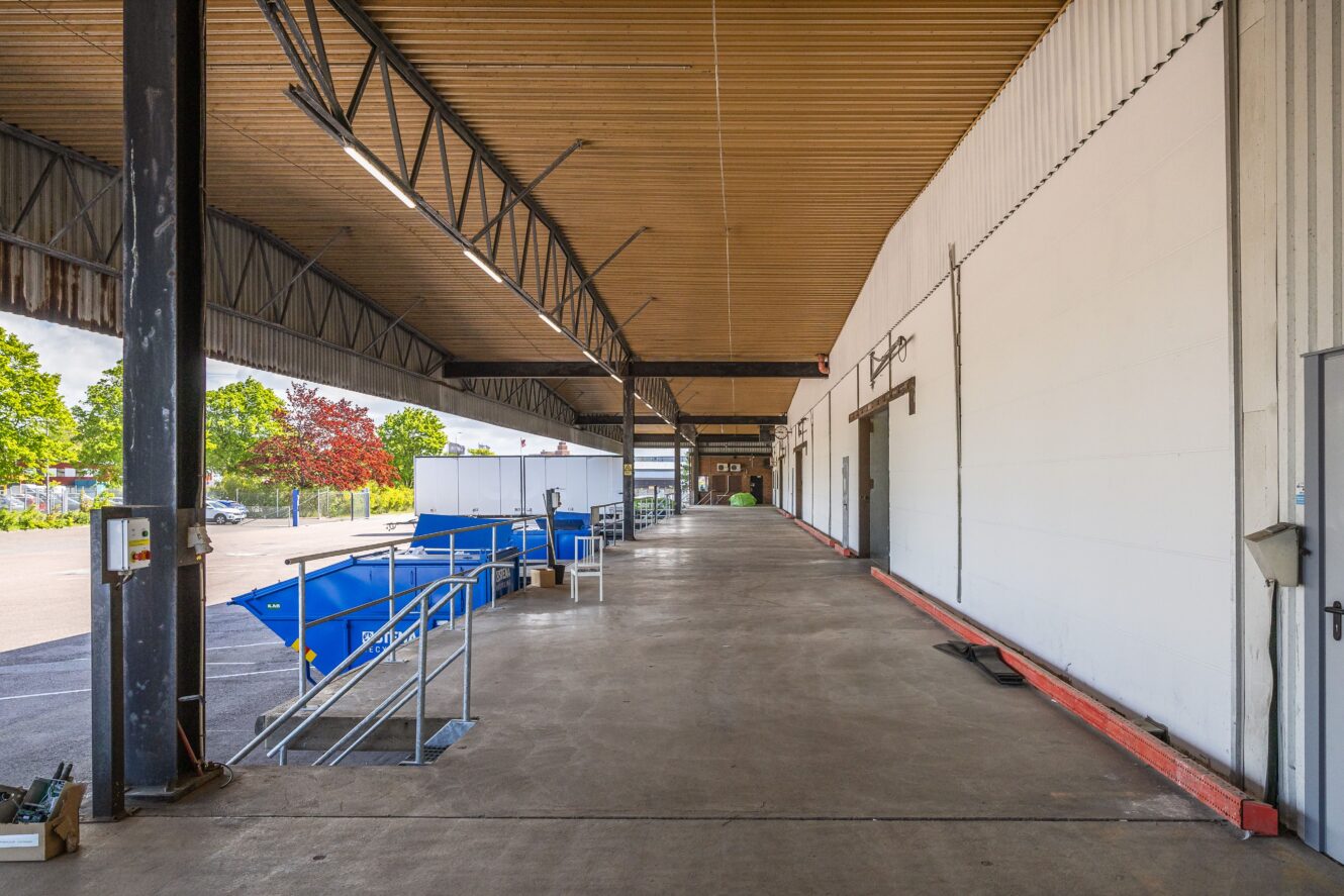 En täckt lastkaj med blåa containrar, metallräcken och takbjälkar intill en stor vit industribyggnad. Träd och himmel syns i bakgrunden.