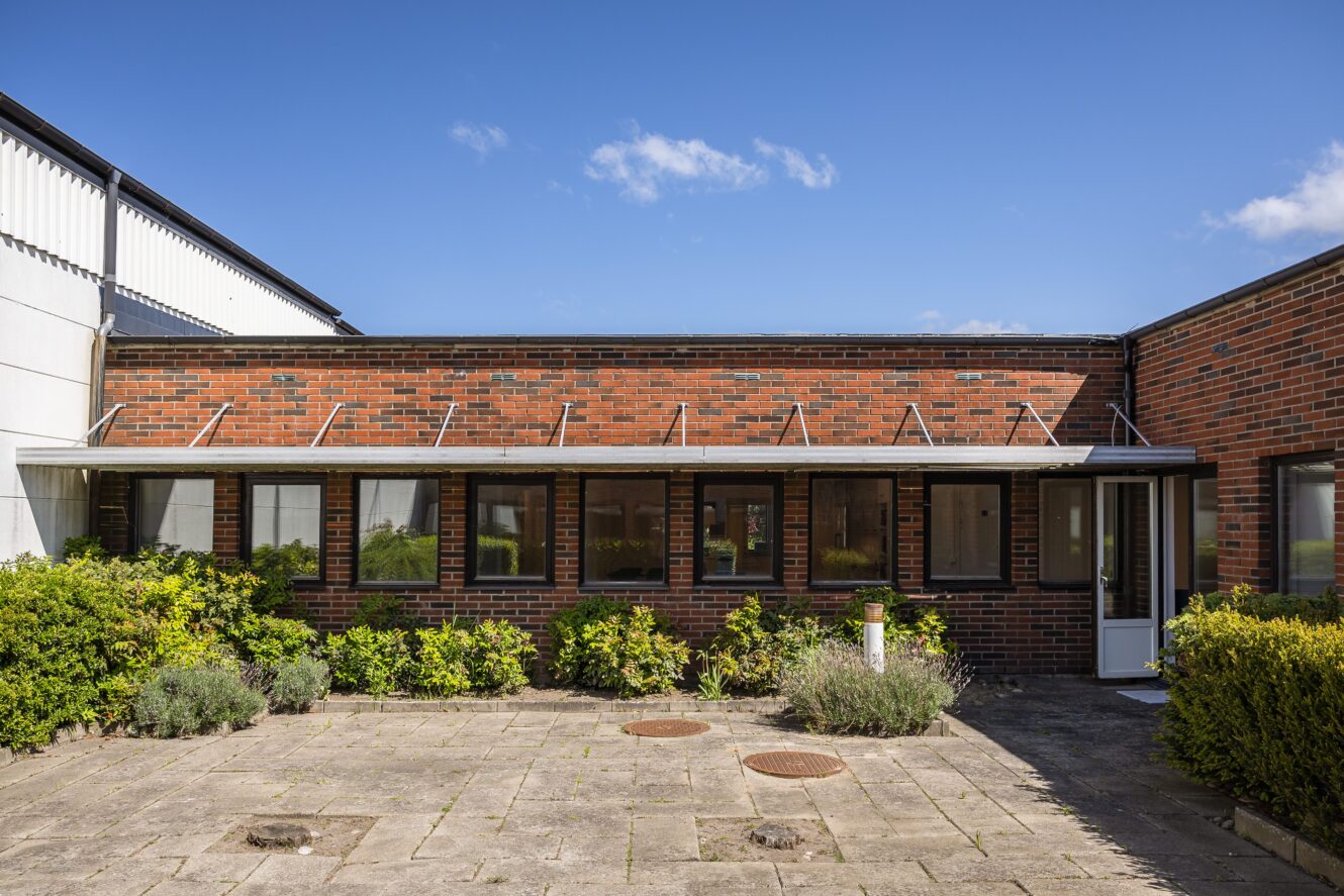 Envånings tegelbyggnad med en rad fönster, ett platt tak och en stenlagd uteplats omgiven av gröna buskar under en klarblå himmel.