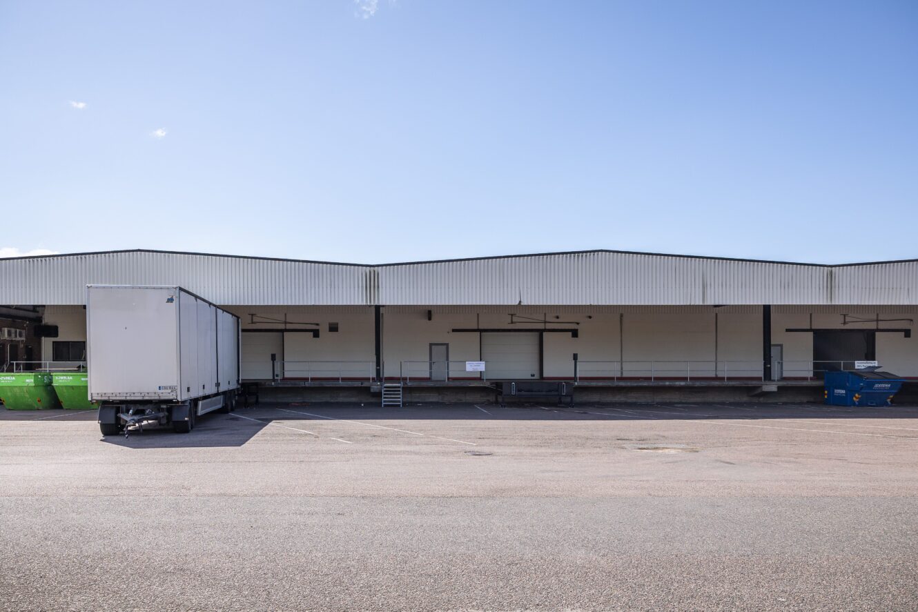 En vit industriell lastkaj med en lastbil med släp parkerad vid ena kajen, blå himmel ovanför och containrar i närheten.