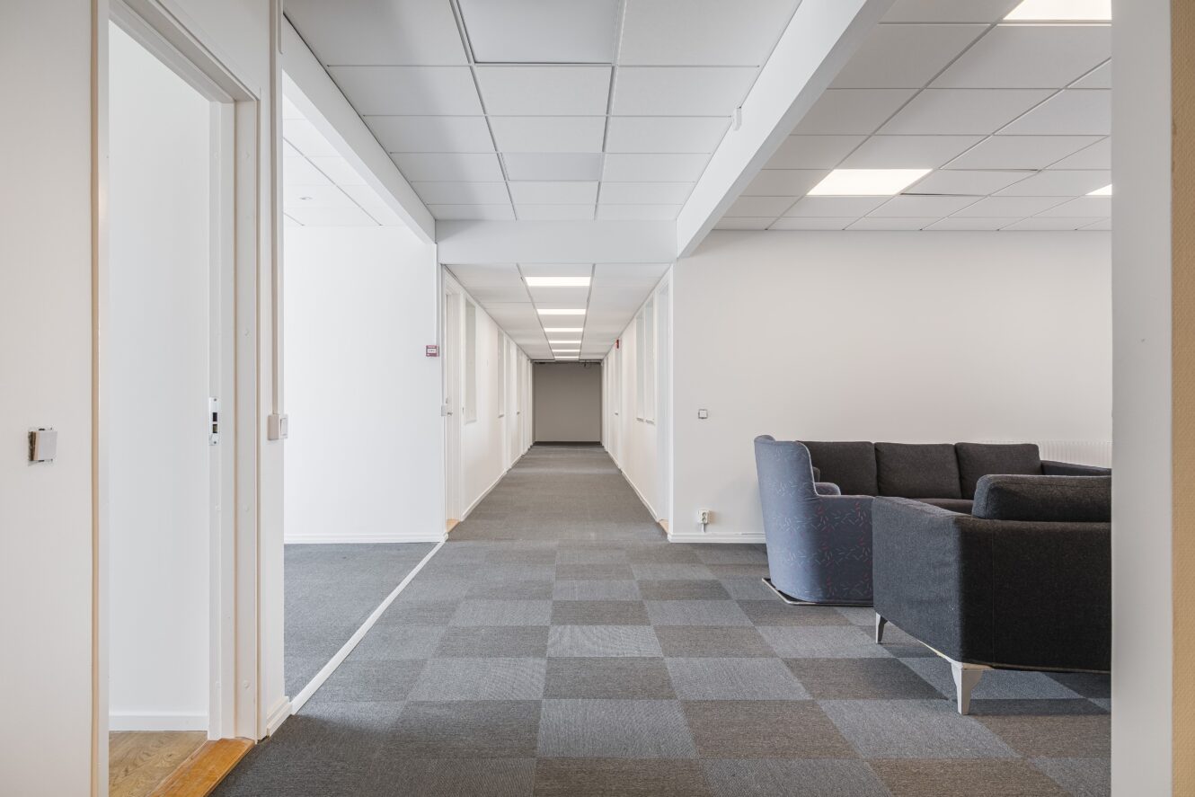 En modern kontorskorridor med grå matta, vita väggar, taklampor och en sittgrupp med mörka soffor på höger sida.