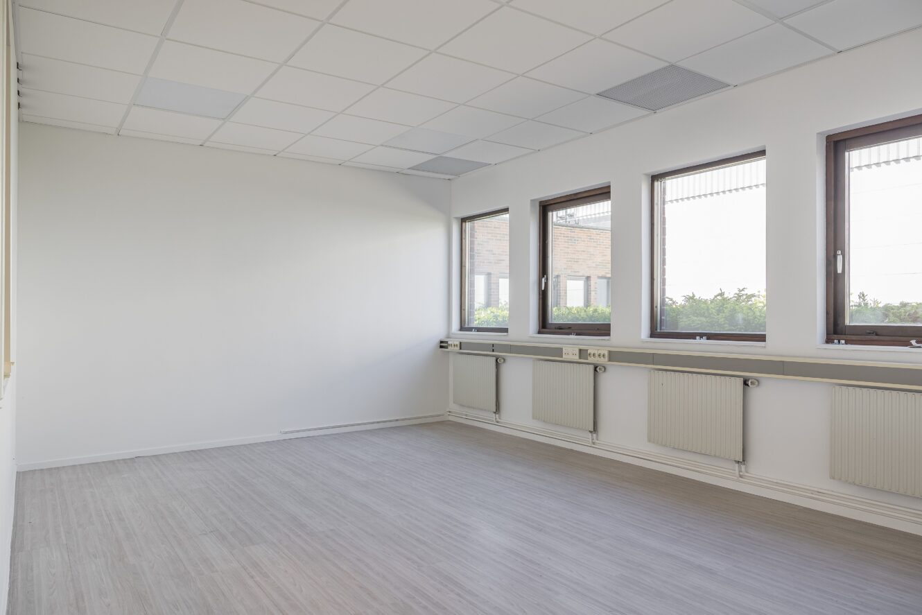 Tomt, omöblerat rum med ljusgrått golv, vita väggar, tre stora fönster och radiatorer under fönstren. Taket har kvadratiska plattor och lysrörsbelysning.