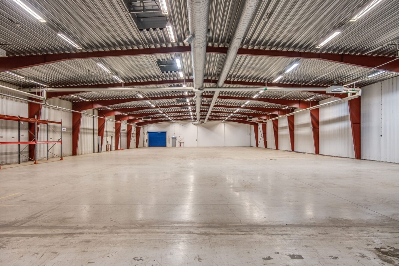 Rymlig, tom industrilokal med betonggolv, synliga takbjälkar, vita väggar och takbelysning. En blå dörr syns i bortre änden.