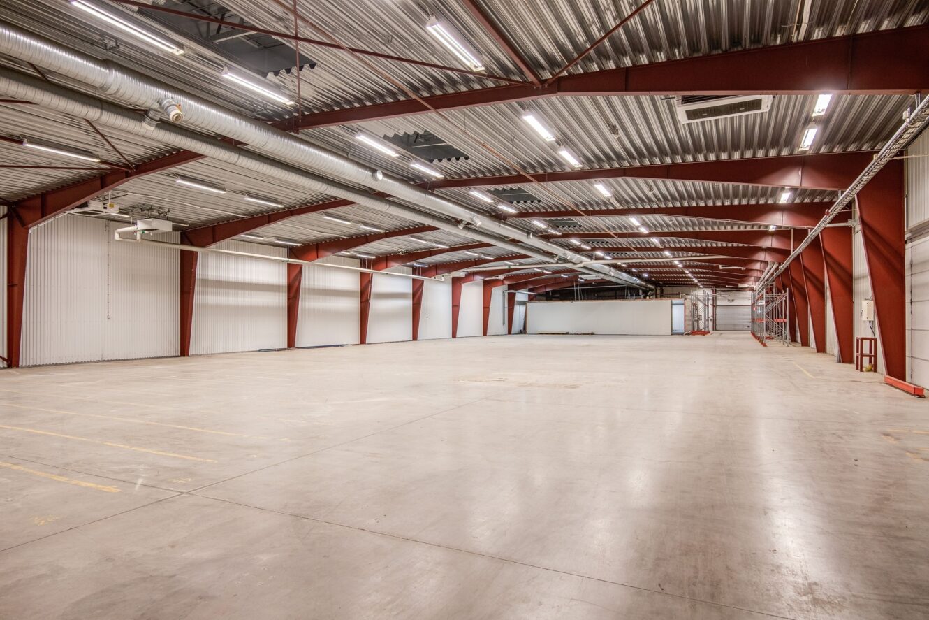 Stor tom lagerlokal med betonggolv, röda stålbalkar och takbelysning. Väggarna är vita och utrymmet är öppet och obebott.