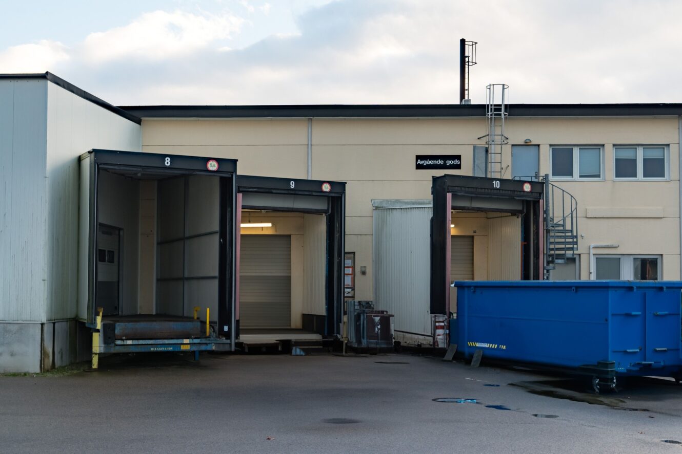 Tre lastbryggor numrerade 8, 9 och 10 vid en lagerbyggnad, med en stor blå container i närheten och en spiraltrappa i metall i bakgrunden.