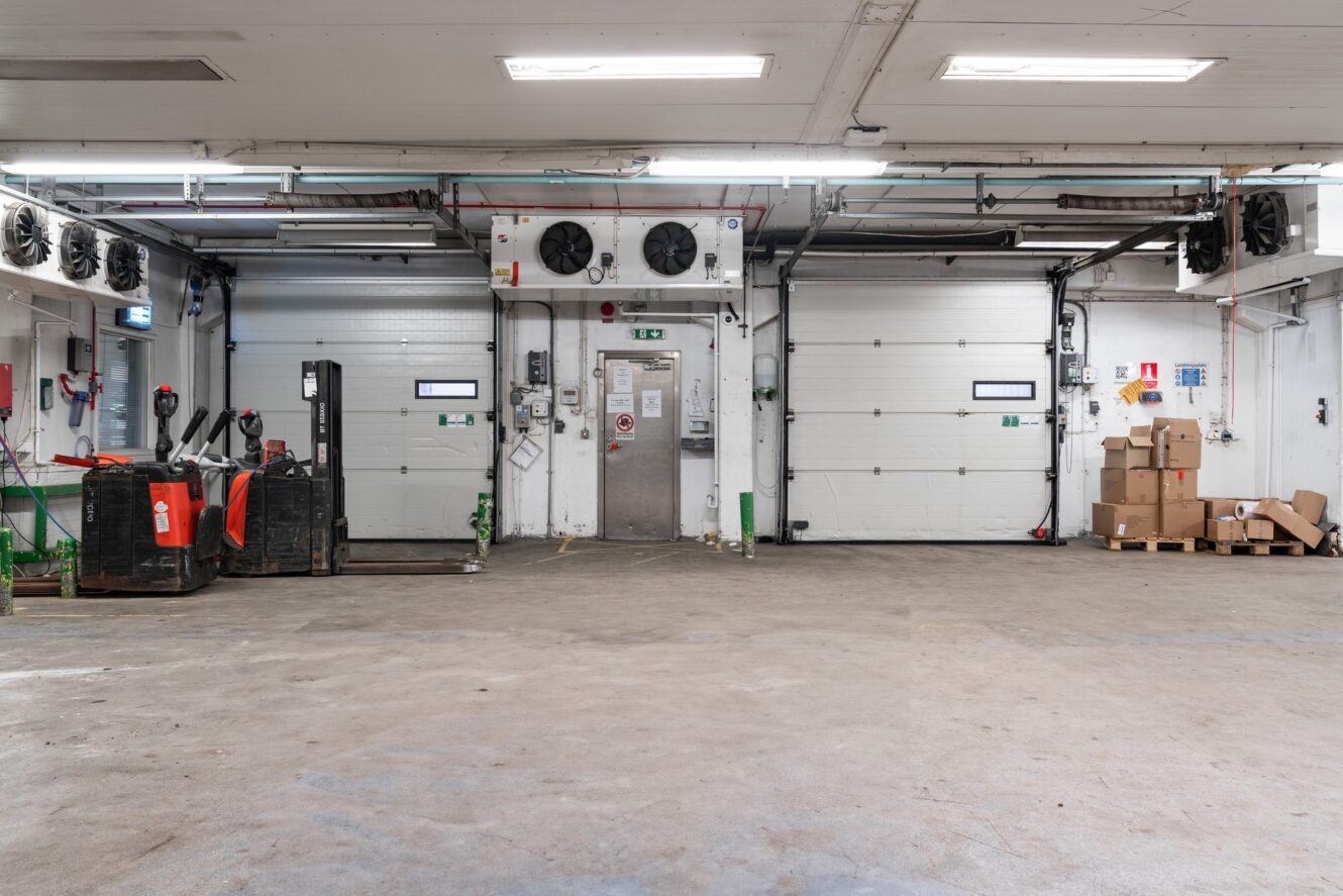 Industriell lagerinredning med betonggolv, pallbockar, kartonger, stora takskjutportar samt väggmonterade fläktar och rör.