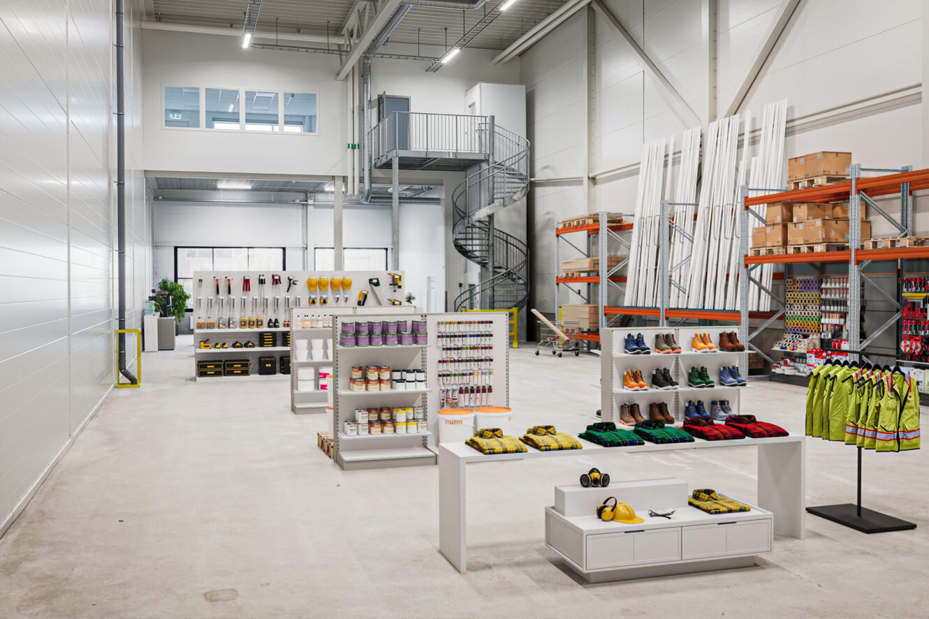 Ett rymligt industrilager med hyllor som visar säkerhetsutrustning, arbetskläder, skor, verktyg och tillbehör; spiraltrappa och förvaringsställ i bakgrunden.