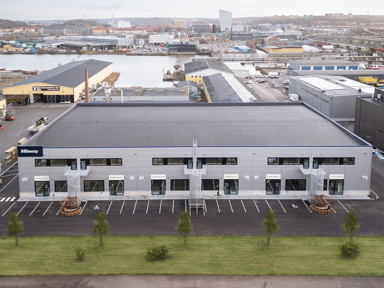 En stor, modern industribyggnad i två våningar med platt tak, flera glasdörrar och intilliggande parkering, belägen i ett industriområde nära vatten.