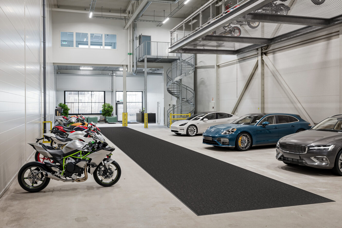Ett modernt inomhusparkeringsgarage med flera lyxbilar och motorcyklar parkerade, med vita väggar, stora fönster och en spiraltrappa.
