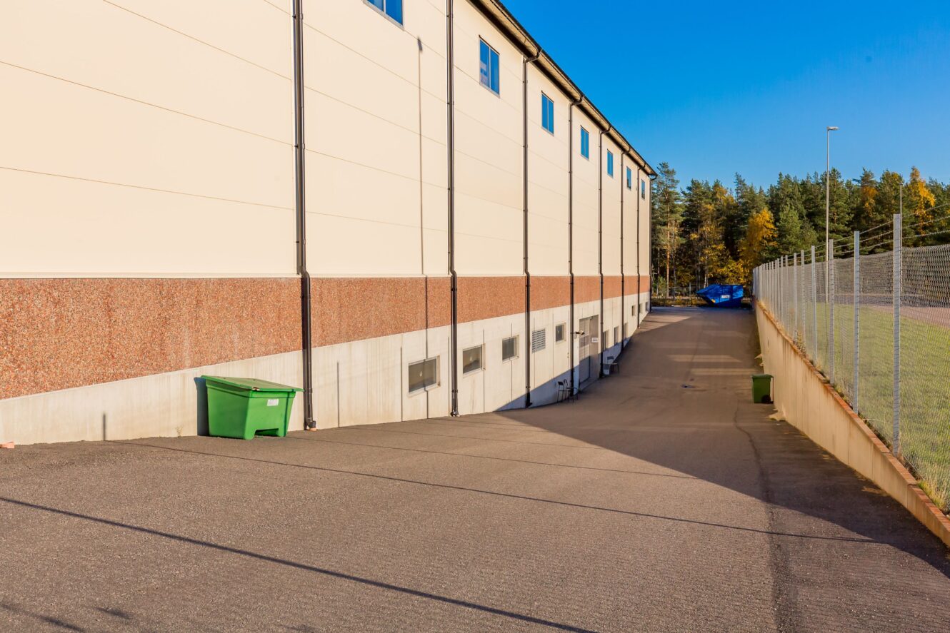 En asfalterad uppfart löper längs en stor byggnad med en grön container nära väggen och ett kedjelänkstaket till höger, under en klarblå himmel.