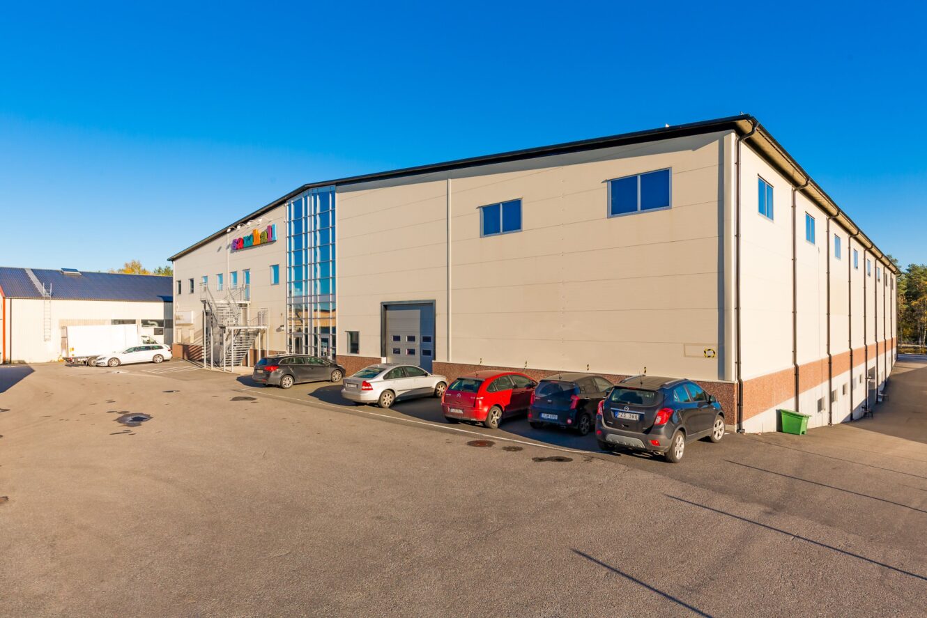 En stor industribyggnad med flera parkerade bilar framför, fotograferad en klar, solig dag.