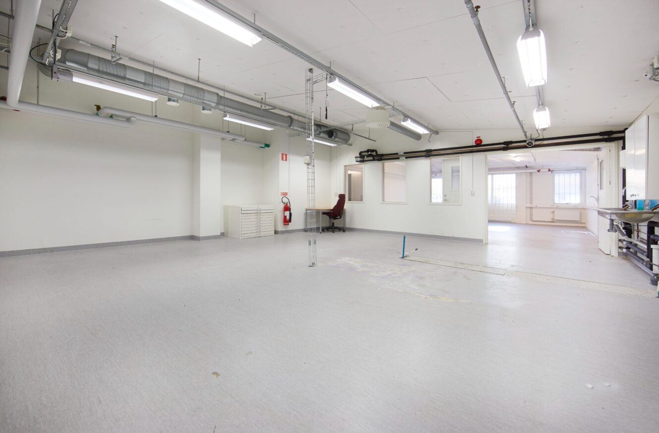 Stort, tomt industrirum med vita väggar, takbelysning, synliga rör, en ensam kontorsstol och en brandsläckare monterad på väggen.