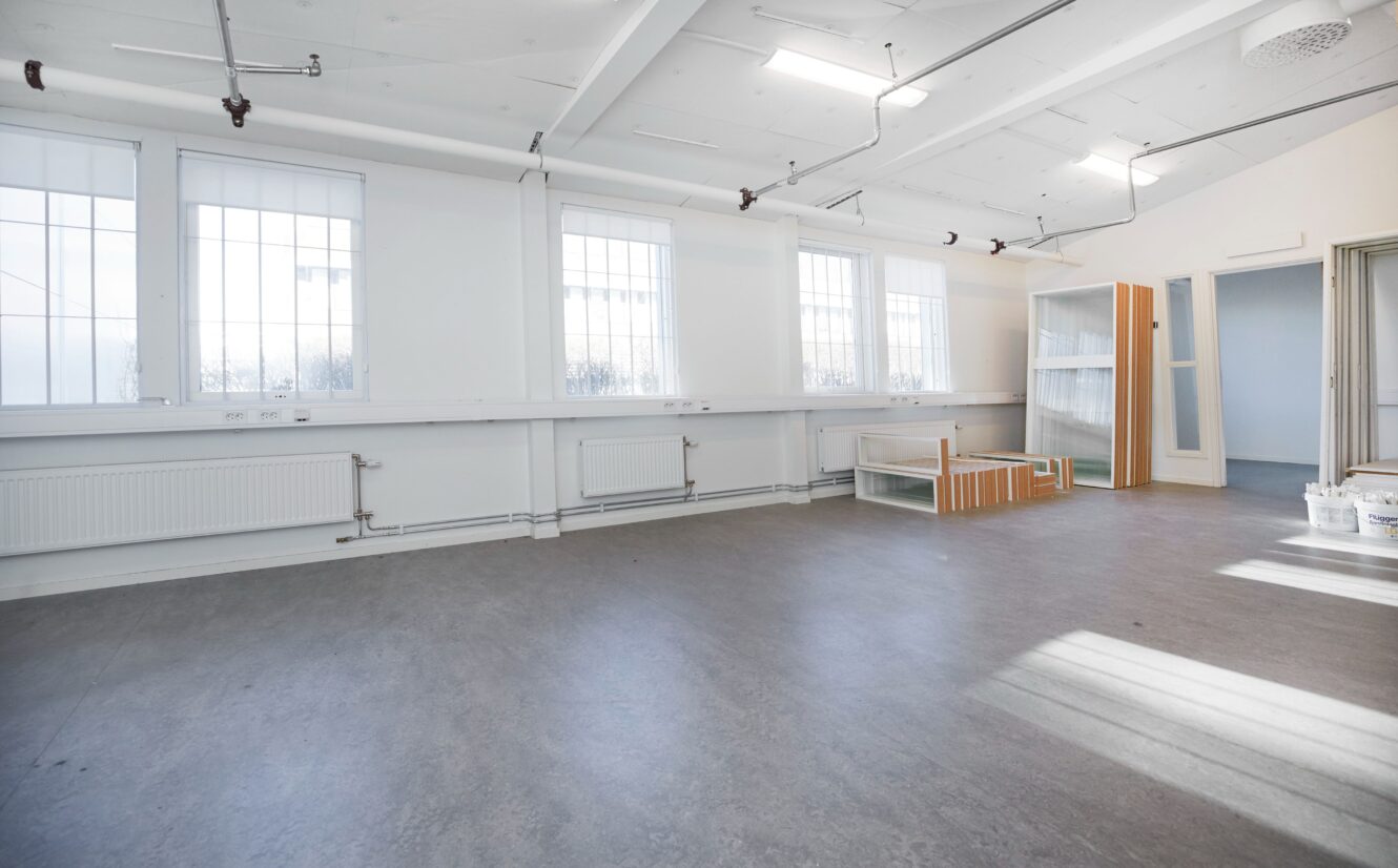Ett tomt, ljust rum med stora fönster, vita väggar, grått golv och några nedmonterade möbler och målarhinkar i ett hörn.