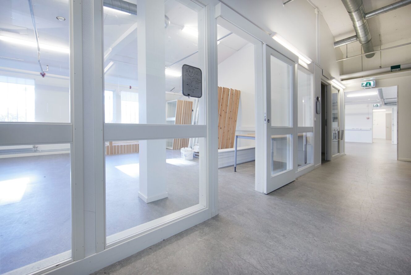 En modern, tom hall med stora fönster och glasdörrar leder till ljusa rum med träbänkar och vita väggar.