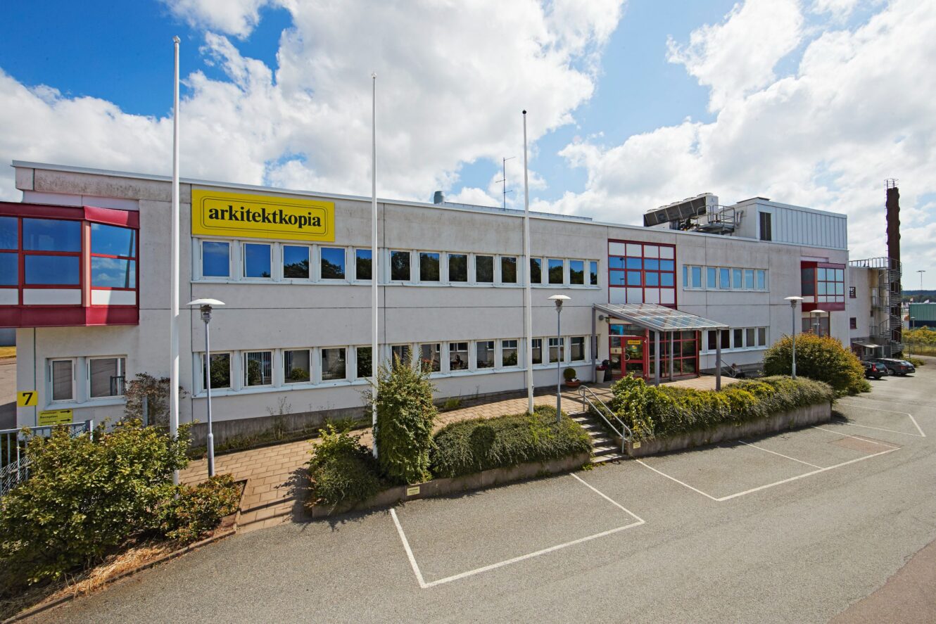 En kontorsbyggnad i två våningar med en skylt med texten arkitektkopia ovanför entrén, flera fönster, tomma parkeringsplatser och en delvis molnig himmel ovanför.