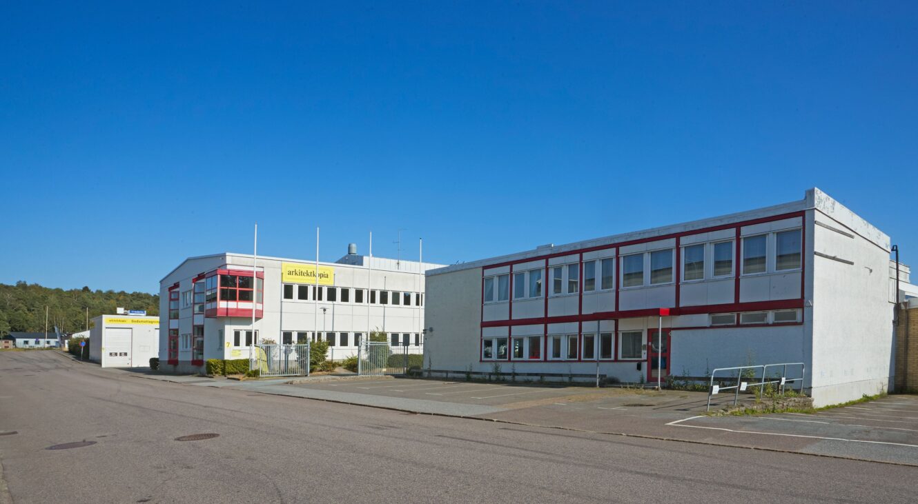 Två rektangulära industribyggnader med vit och röd exteriör står intill en bred, tom gata under en klarblå himmel.