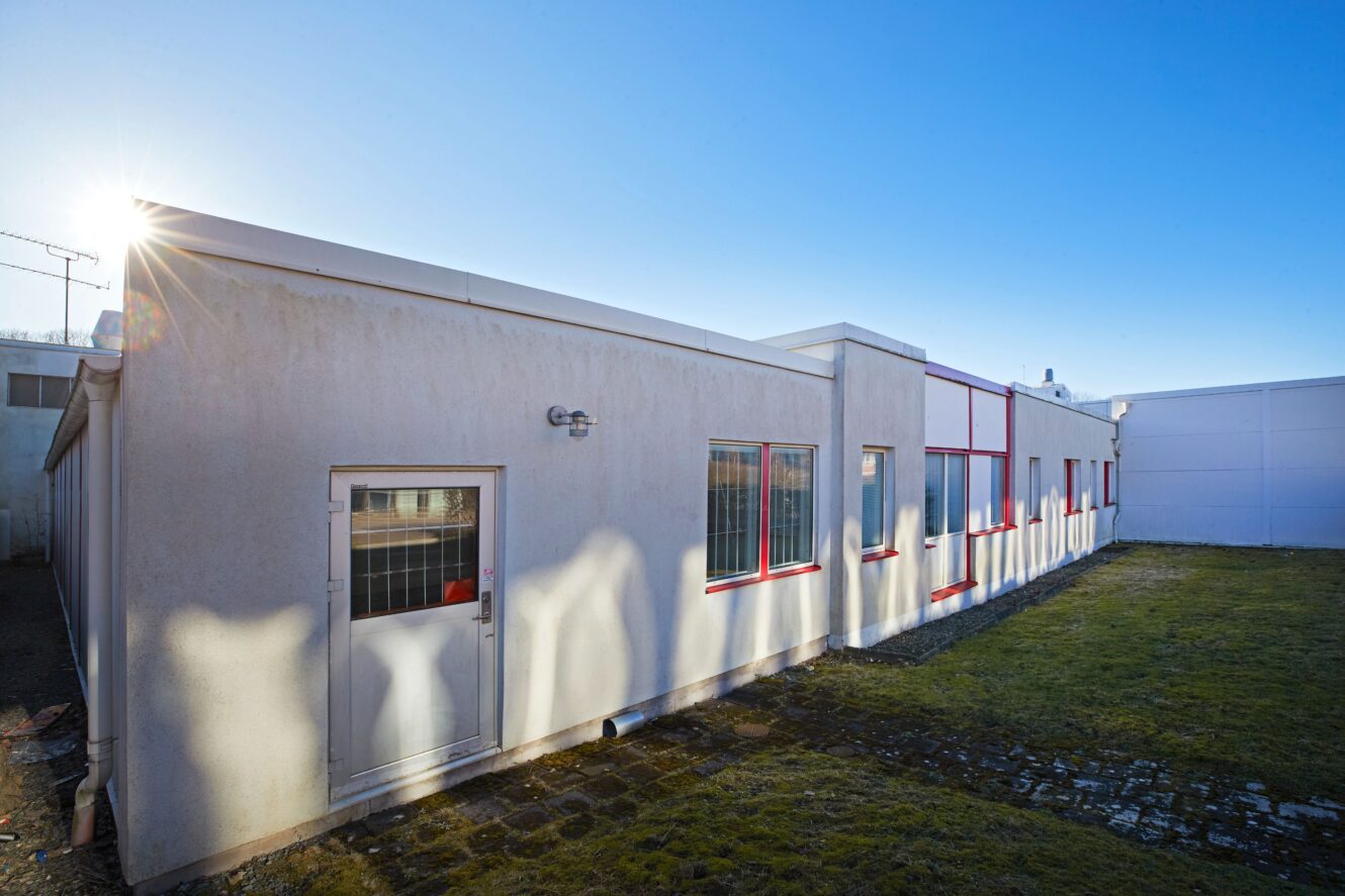 En vit envåningsbyggnad med röda fönster och dörrar, som kastar långa skuggor på väggen, står på en gräsmatta under en klarblå himmel.