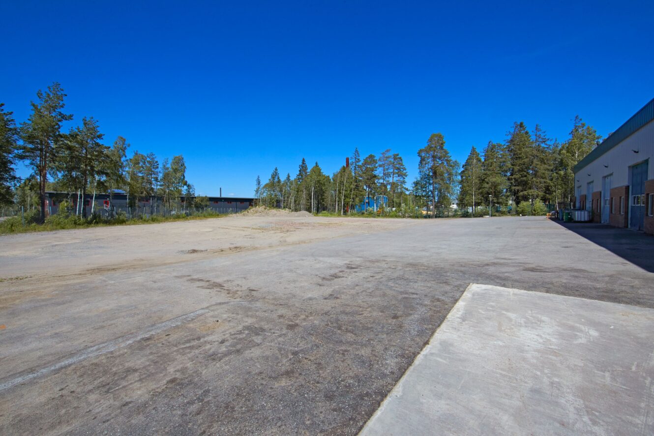 En stor, tom betongtomt som kantas av träd under en klarblå himmel, med en byggnad till höger i bild.