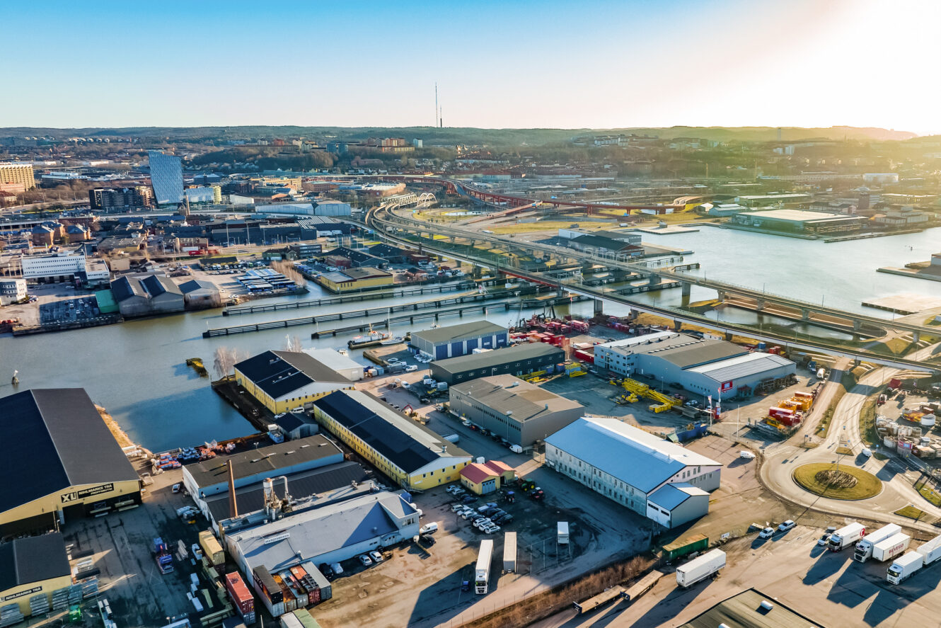 Flygfoto över ett industriellt hamnområde med lagerbyggnader, vägar, broar och vattenkanaler under en klar himmel.
