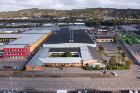 Ett stort industribyggnadskomplex med en blandning av tegel- och metallkonstruktioner, parkeringsplatser och träd, beläget nära en väg och omgivet av andra industrianläggningar och kullar.