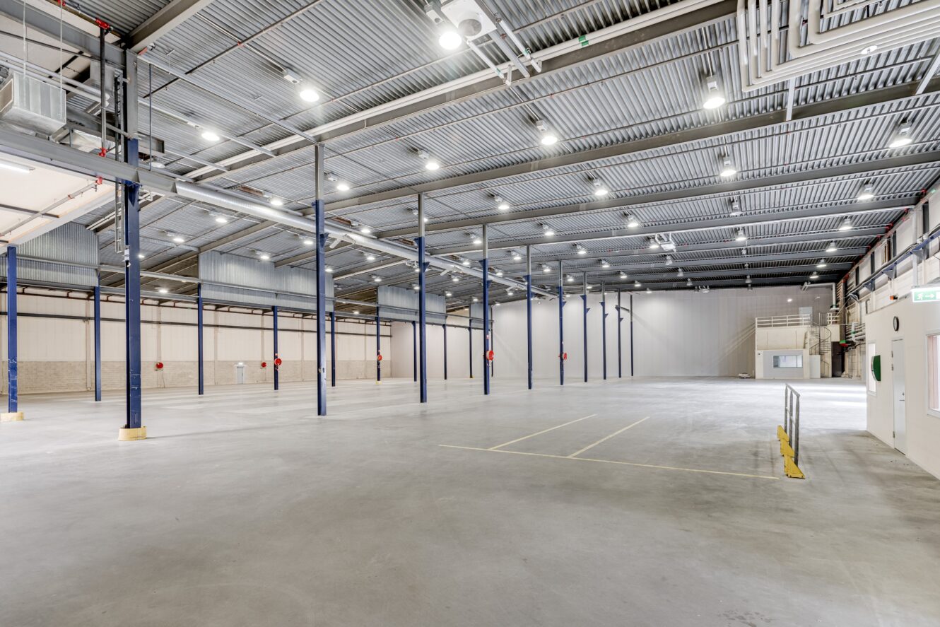 Interiör i en stor, tom lagerlokal med betonggolv, högt i tak, stålbalkar, stark belysning och synliga rör.