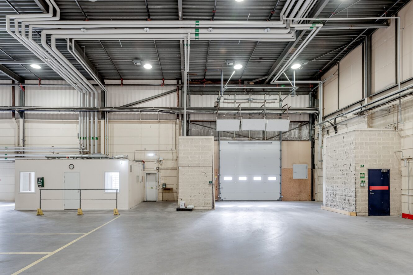 Tomt industriellt lagerutrymme med betonggolv, synliga rör i taket, en stor garageport och ett litet slutet kontorsutrymme till vänster.