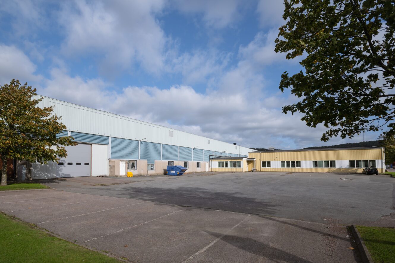 Stort industriellt lager med blå och vit exteriör, flera garageportar, en gul lastkaj och en tom parkeringsplats under en delvis molnig himmel.