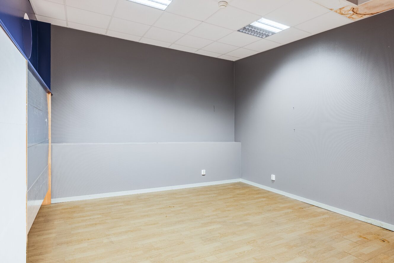 Tomt rum med ljust trägolv, grå väggar, vitt tak med lysrör och eluttag på väggarna.