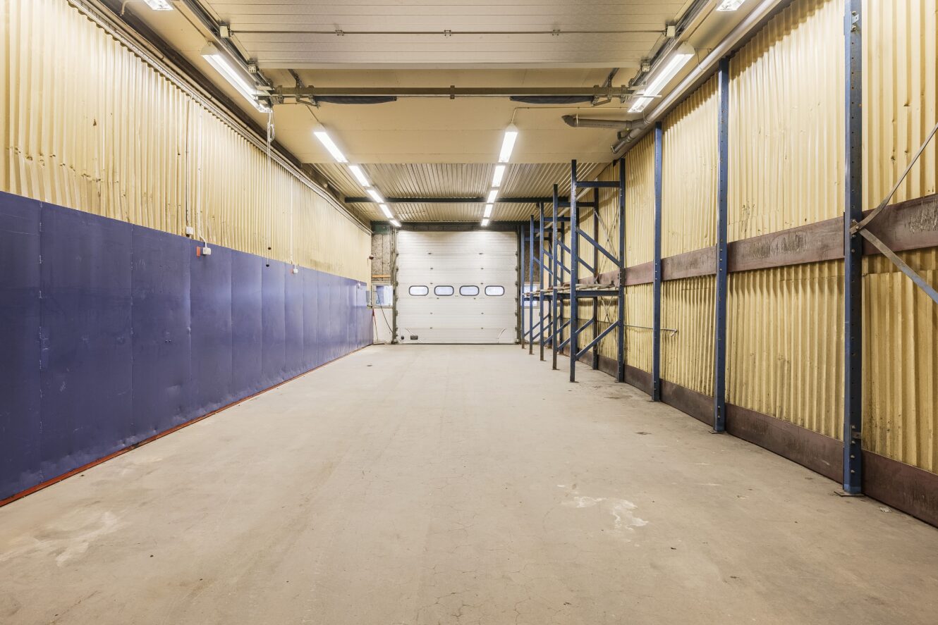 Tomt industrilager med gula väggar, blå nedre panel, taklampor, metallhyllor till höger och en stängd vit garageport längst bort.