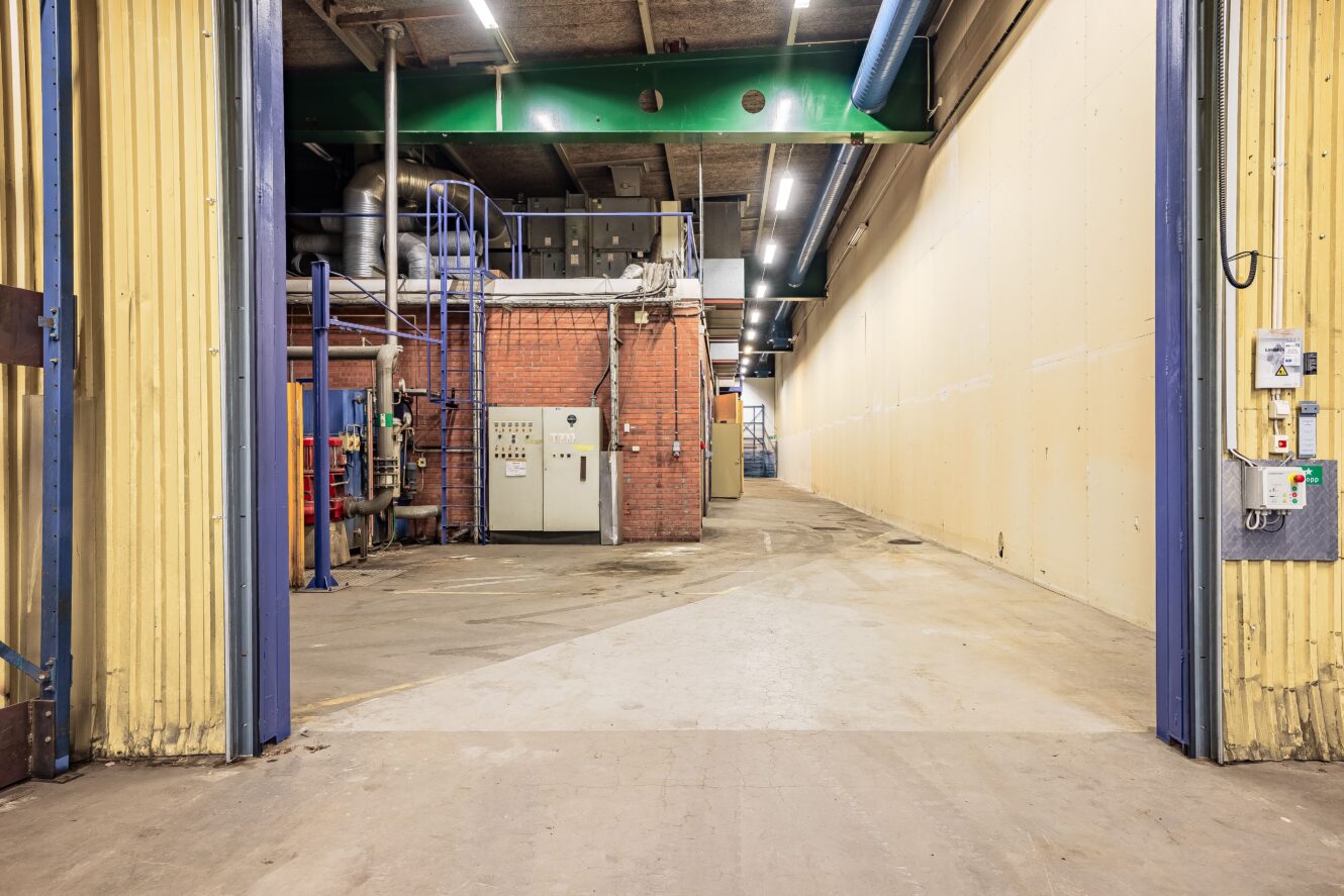 En rymlig industriell lagerlokal med exponerade tegelmaskiner, kontrollpaneler, rör och takbelysning, sett från en öppen entré.