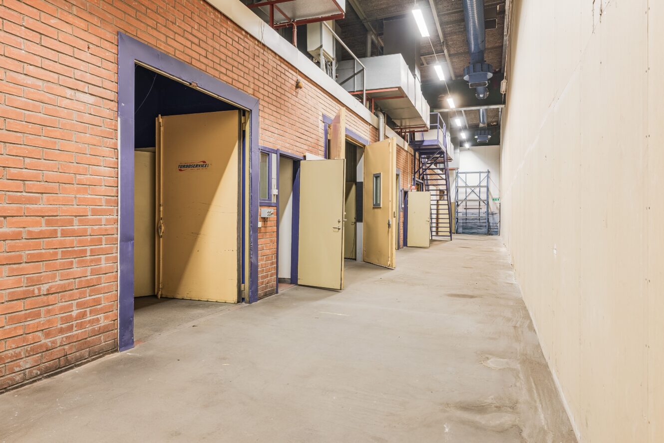 En bred korridor med tegel och släta väggar, flera öppna dörrar som leder till små rum och en metalltrappa längst bort.