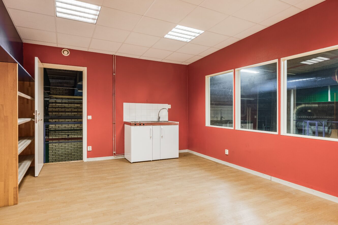 Ett litet rum med röda väggar, ljust trägolv, vitt tak, stora innerfönster, ett vitt diskbänksskåp och trähyllor vid en öppen dörr.