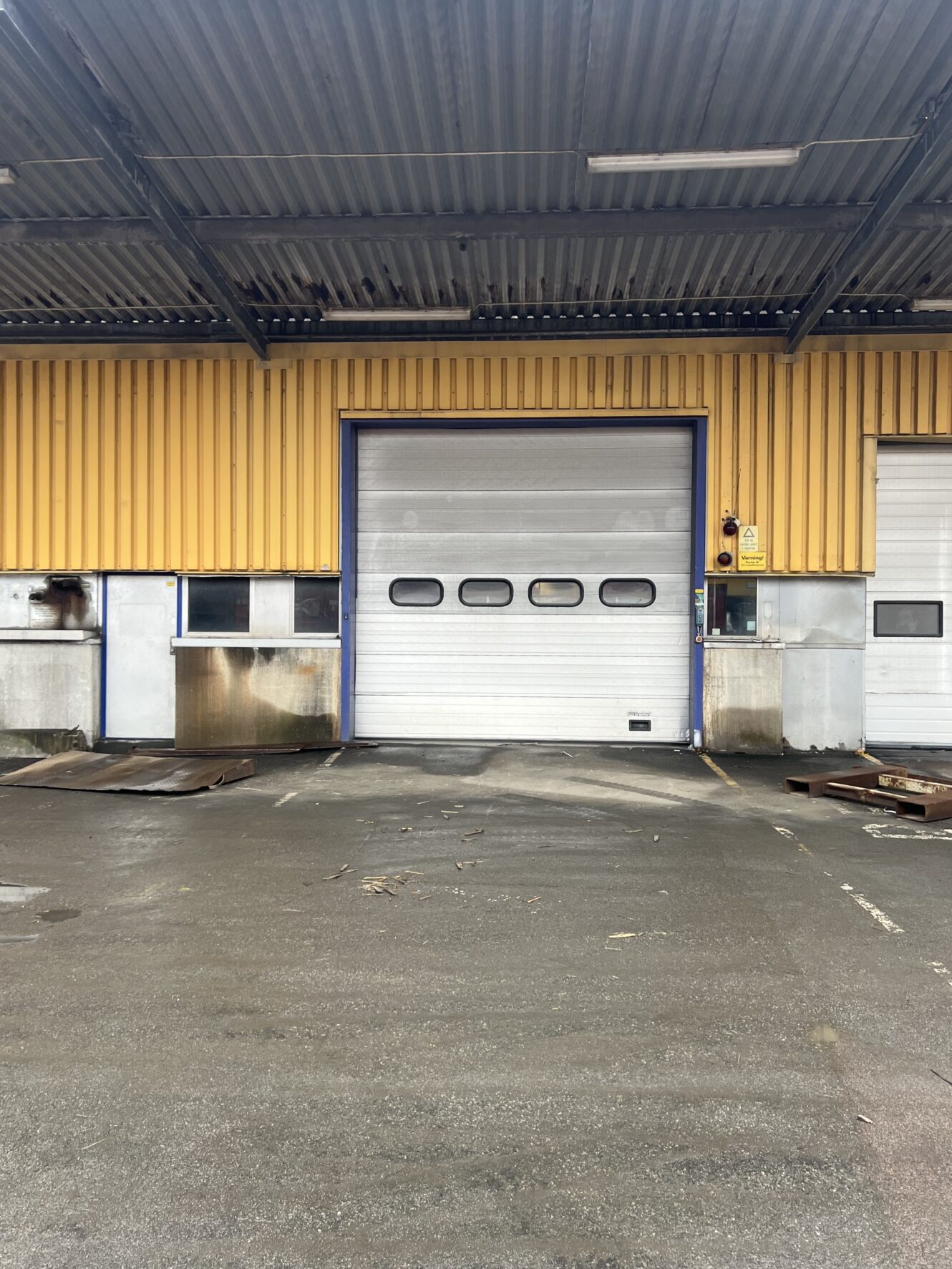 En stängd industriell garageport i en gul metallbyggnad, med våt trottoar och lite skräp framför.