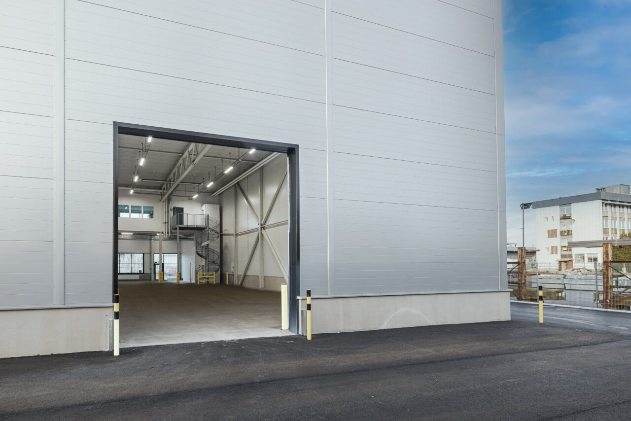 En stor öppen garageport på ett modernt industriellt lager med en asfalterad uppfart, invändig belysning och utsikt över närliggande byggnader.