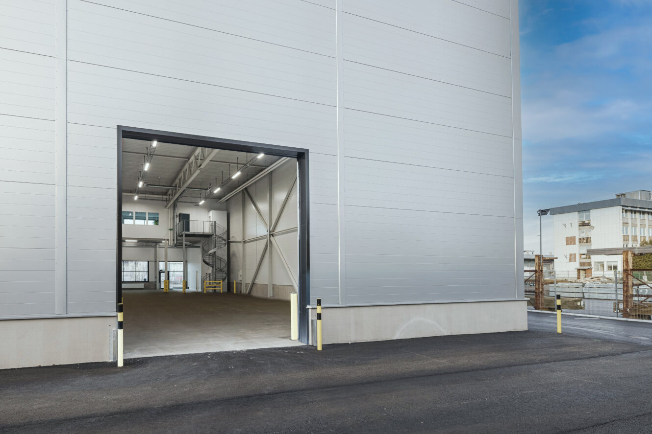 En stor öppen garageport på ett modernt industriellt lager med en asfalterad uppfart, invändig belysning och utsikt över närliggande byggnader.