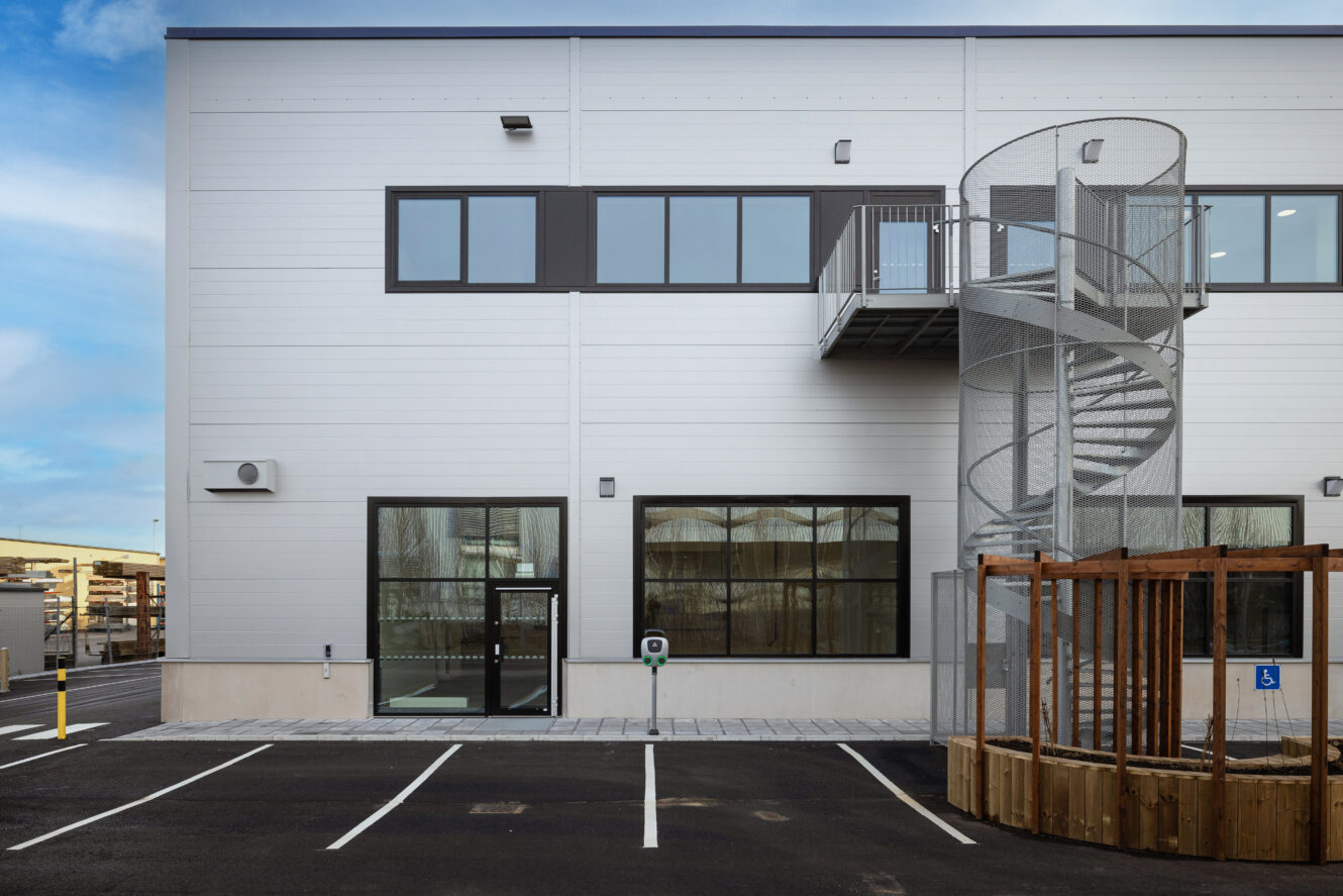 Modern industribyggnad med stora fönster, ingång i glas, spiraltrappa på utsidan, tomma parkeringsplatser och en inhägnad i trä på en asfalterad tomt.