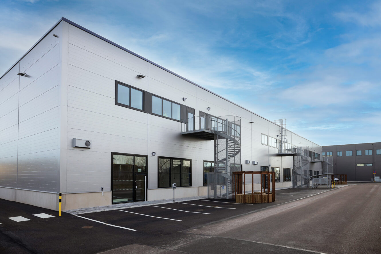 Modern industribyggnad med metallfasader, utvändiga trappor, stora fönster och en intilliggande parkeringsplats under en delvis molnig himmel.