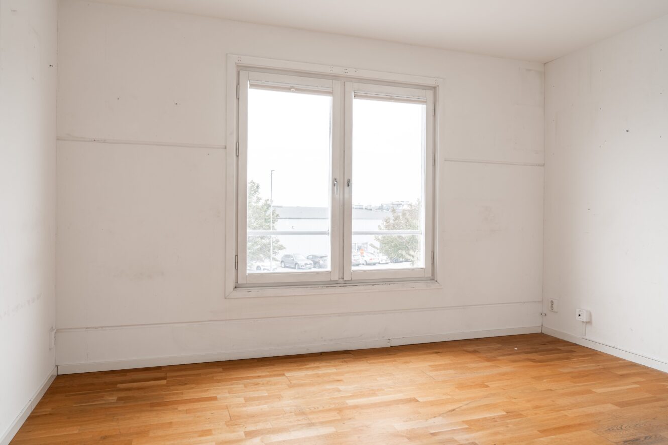 Tomt rum med vita väggar, ett stort fönster och ljust trägolv. Utsikten utanför visar träd, en parkeringsplats och en byggnad i fjärran.