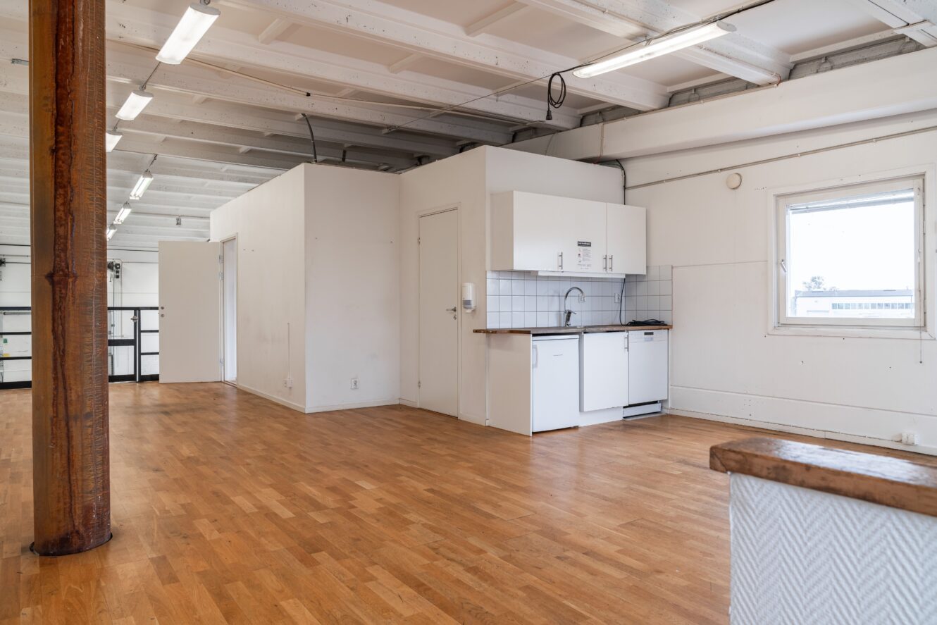 Rymligt rum med trägolv, vita väggar, ett litet pentry, synliga takbjälkar och ett stort fönster som släpper in naturligt ljus.