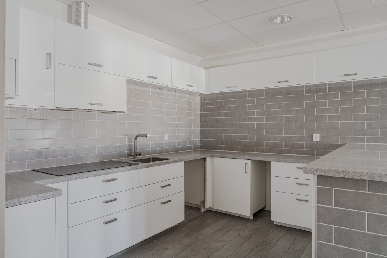 Modernt kök med vita skåp, grå bänkskivor och grått kaklat stänkskydd. Köket ser oavslutat ut med några lådor och skåpsluckor som saknas.