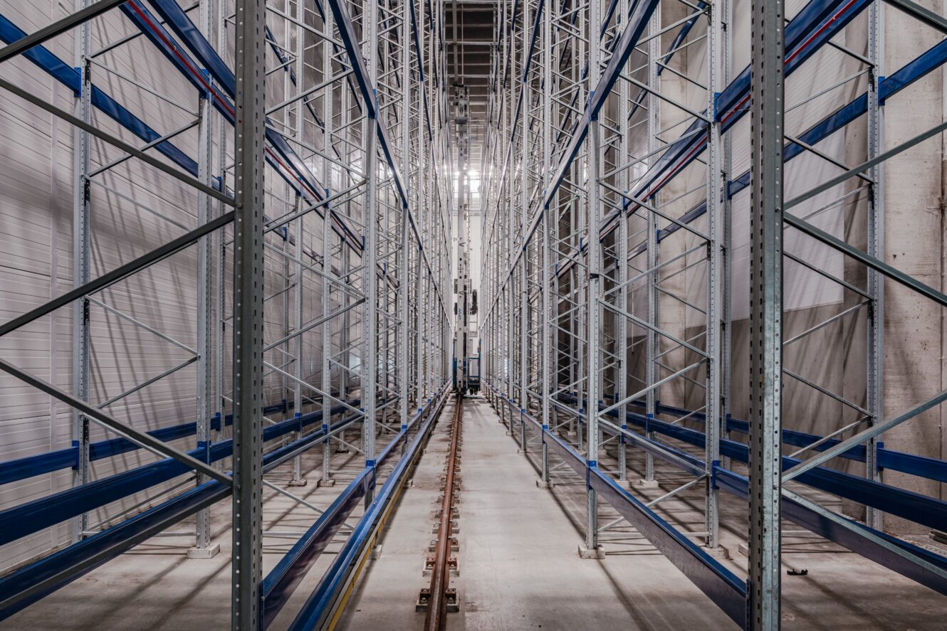 Tomma industriella lagerhyllor med metallställ och järnvägsspår som löper längs mitten, sett från ett centralt perspektiv.