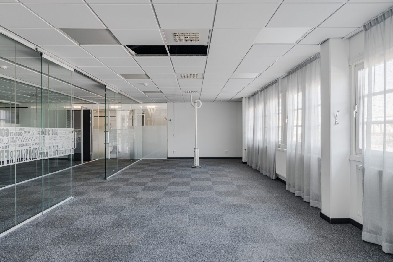 Tomt modernt kontorsutrymme med glasväggar, grå heltäckningsmatta och stora fönster med skira gardiner; vissa takplattor saknas eller har flyttats.