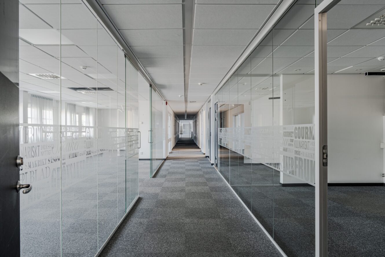 En modern kontorskorridor med glasväggar, heltäckningsmatta och flera dörrar som leder till tomma rum, upplyst av naturligt och artificiellt ljus.