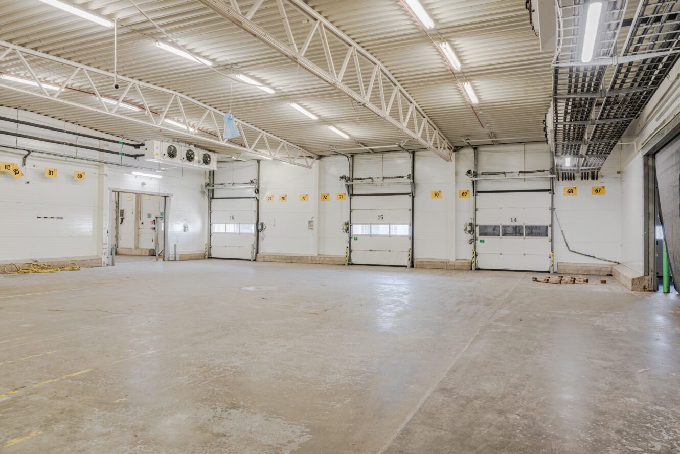 Rymlig, tom industrilagerlokal med betonggolv, högt i tak, synliga bjälkar och flera stora garageportar längs ena väggen.