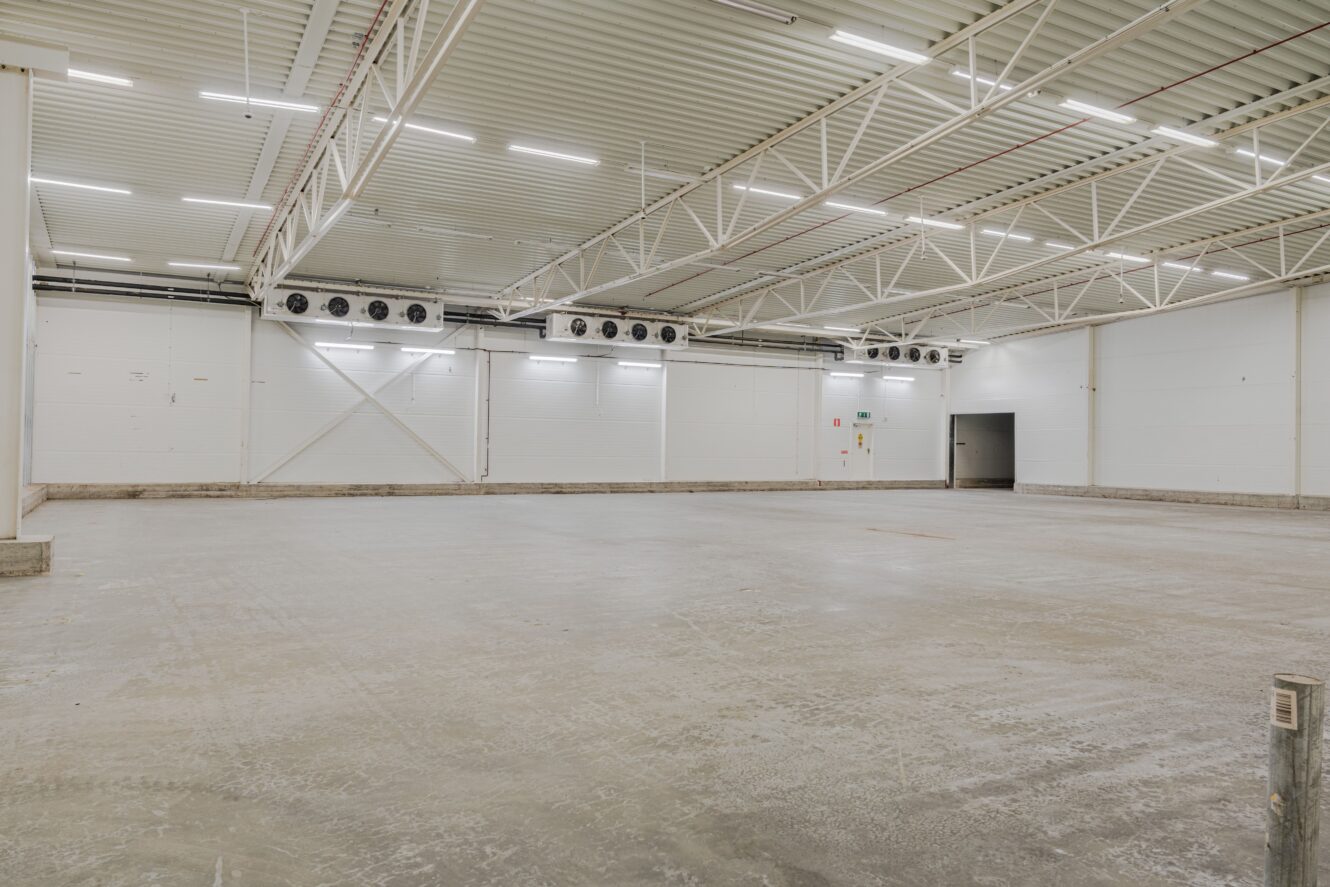 Tomt industrilager med betonggolv, vita väggar, synliga takbjälkar, ventilationsaggregat och takbelysning.