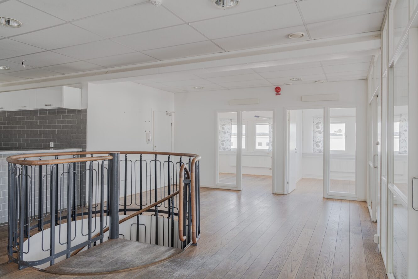 Ljus, modern interiör med trägolv, spiraltrappa, glasdörrar och vita väggar; köksdel med bakplatta i grått kakel syns till vänster.