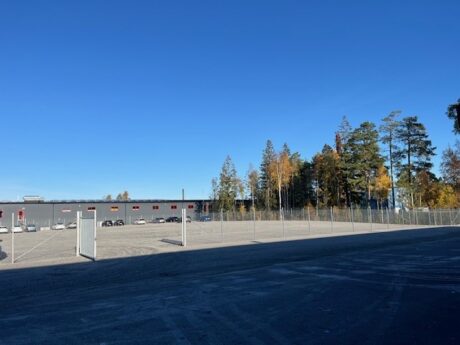 En inhägnad, tom grusplan med en öppen grind, träd med höstlöv och en grå industribyggnad i bakgrunden under en klarblå himmel.