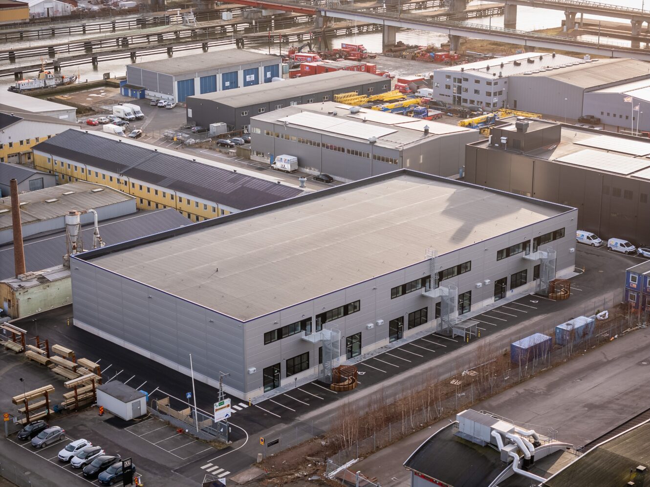 Flygfoto över ett stort industrilager med parkeringsplats, omgivet av andra kommersiella byggnader och vägar i ett industriområde.