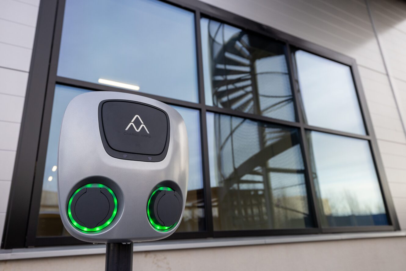 Närbild av en laddningsstation för elfordon med två portar, placerad framför en byggnad med stora fönster som speglar en spiraltrappa.
