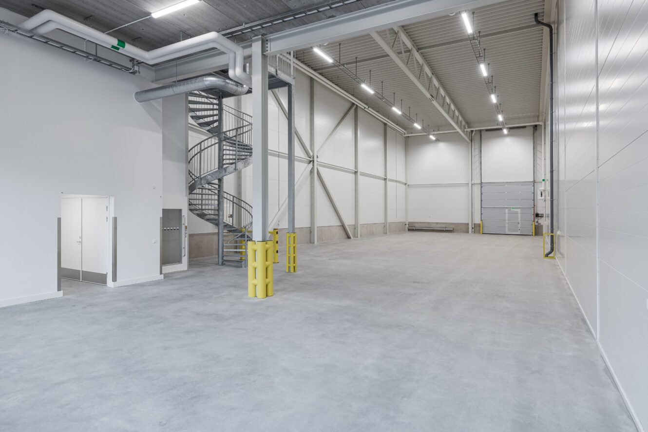 Tomt industrilager med betonggolv, högt i tak, vita väggar, spiraltrappa i metall, stor takskjutport och lysrörsbelysning. Gula säkerhetsbarriärer finns på plats.