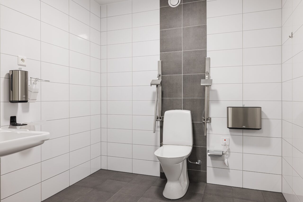 Tillgänglig offentlig toalett med vitkaklade väggar, toalett med stödskenor, handfat, tvålpump, pappershanddukspump och nödstopp.